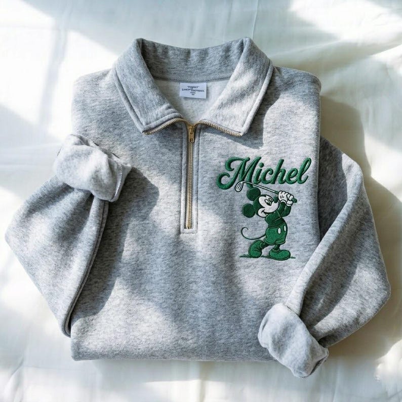 May include: A gray sweatshirt with a collared neckline and a gold zipper. The sweatshirt features an embroidered design of Mickey Mouse playing golf, with the name "Michel" in green script.