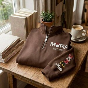 May include: A brown quarter-zip sweatshirt with the word "MOMA" and a Minnie Mouse graphic, along with "EST.2026" printed on the front. The sleeve features a Minnie Mouse design and the name "Mia". The sweatshirt is displayed on a wooden table.
