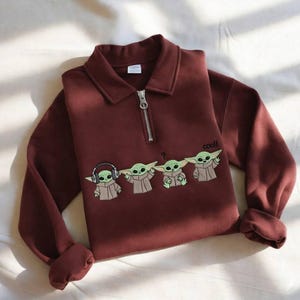 May include: A maroon long-sleeve sweatshirt with a collared neckline and a silver zipper. The sweatshirt features an embroidered design of four Grogu characters, with the word "cool!" above the last character.