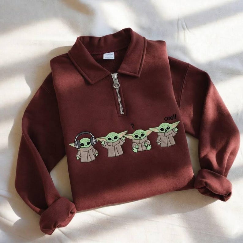 May include: A maroon long-sleeve sweatshirt with a collared neckline and a silver zipper. The sweatshirt features an embroidered design of four Grogu characters, with the word "cool!" above the last character.