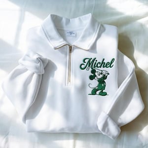 May include: White sweatshirt with a collared neckline and a gold zipper. The sweatshirt features a green embroidered design of Mickey Mouse playing golf, with the name "Michel" above the character.