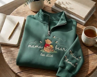 May include: A green quarter-zip sweatshirt with embroidered Winnie the Pooh design. The design includes the words "mama bear" and "Est. 2026". The sleeve has the names "Mina" and "Thomas" embroidered with bees. The sweatshirt is on a wooden table.