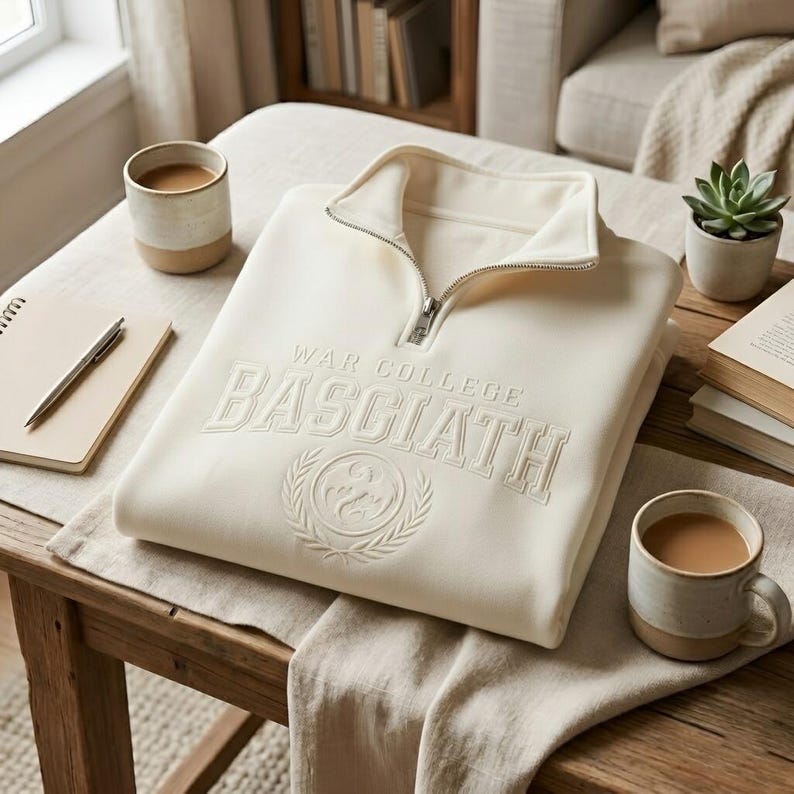 Puede incluir: Sudadera crema con cuello con cremallera de cuarto, con el texto "WAR COLLEGE BASCIATH" en relieve. Un emblema circular con una corona de laurel est&aacute; debajo del texto. La sudadera est&aacute; doblada sobre una mesa de madera, con tazas de caf&eacute; y una planta suculenta cerca.