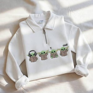 May include: White sweatshirt with a collared neckline and a silver zipper. The sweatshirt features an embroidered design of four Grogu characters, with the word "cool!" printed next to the last character.