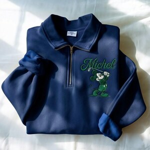 May include: Navy blue golf-themed sweatshirt with a quarter-zip closure and a collared neckline. Features an embroidered Mickey Mouse graphic in green and white, swinging a golf club, with the name "Michel" in green script.