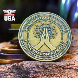 May include: A bronze-colored coin with the text "PRAY WITHOUT CEASING" and "1 THESSALONIANS 5:17" encircling a design of praying hands and a tree. The coin is set on a textured rock surface, with "MADE IN THE USA" text in the upper left.