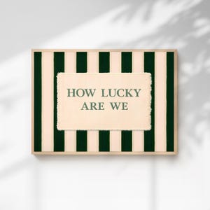 May include: A framed art print with a striped background of alternating dark green and cream vertical stripes. The center features a rectangular, textured patch with the text "HOW LUCKY ARE WE" in green.