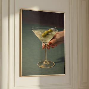 May include: A framed print of a martini cocktail with three olives on a skewer. The clear drink is in a classic stemmed glass, held by a hand with red nail polish. The background is a dark green and burgundy.