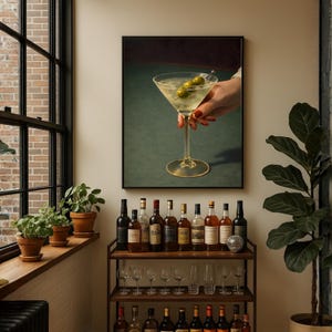 Op de afbeelding: Een ingelijste print van een martini glas met olijven, vastgehouden door een hand met rode nagellak. De print wordt weergegeven boven een bartrol met flessen sterke drank en glaswerk. De achtergrond is een groen oppervlak.