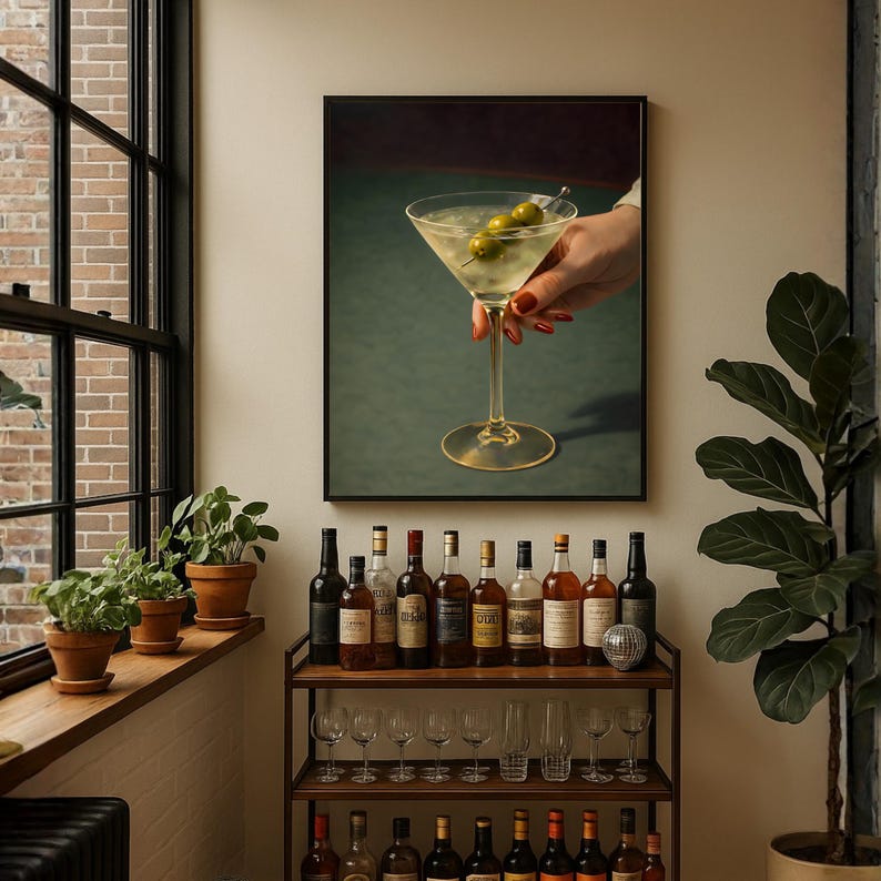 Op de afbeelding: Een ingelijste print van een martini glas met olijven, vastgehouden door een hand met rode nagellak. De print wordt weergegeven boven een bartrol met flessen sterke drank en glaswerk. De achtergrond is een groen oppervlak.
