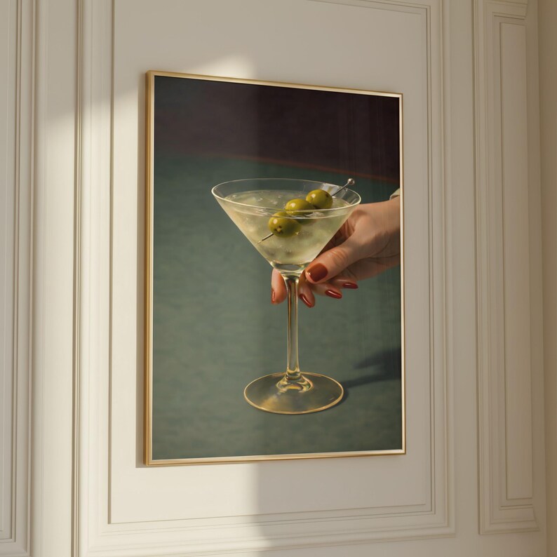 May include: A framed print of a martini cocktail with three olives on a skewer. The clear drink is in a classic stemmed glass, held by a hand with red nail polish. The background is a dark green and burgundy.