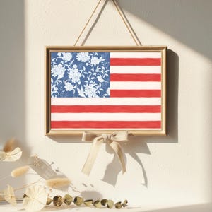 May include: A rectangular wall hanging featuring a stylized American flag design. The canton displays a blue background with white floral and bird patterns, while the field consists of horizontal red and white stripes. A beige ribbon bow is attached.