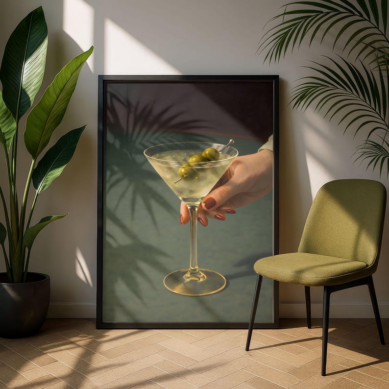 Op de afbeelding: Een ingelijste print van een martini-cocktail met drie olijven aan een spies. De martini zit in een klassiek glas, vastgehouden door een persoon met rode nagellak. De achtergrond is een gedempt groen met schaduwen van palmbladeren.