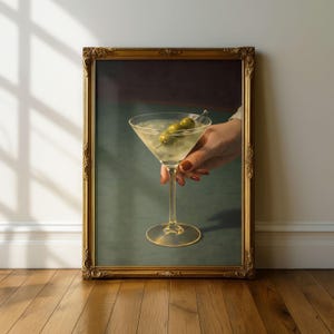 Op de afbeelding: Een ingelijste print van een martini-cocktail met olijven. Het heldere glas wordt vastgehouden door een hand met rode nagellak. De gouden lijst heeft sierlijke details, tegen een groene achtergrond.