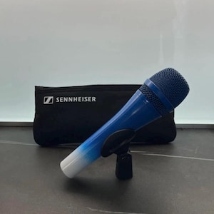 Puede incluir: Un micrófono azul y blanco sobre un soporte negro. El micrófono tiene una cabeza de malla azul y un cuerpo degradado de azul a blanco. Una funda negra con el logotipo de Sennheiser está detrás del micrófono.