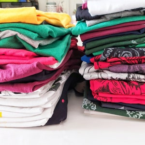 May include: A stack of folded clothing in various colors, including yellow, green, pink, red, and white. The clothing is neatly folded and stacked on a white surface.