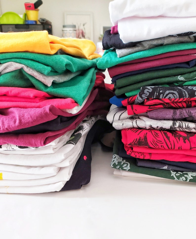 May include: A stack of folded clothing in various colors, including yellow, green, pink, red, and white. The clothing is neatly folded and stacked on a white surface.