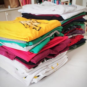May include: A stack of folded t-shirts in various colors, including white, yellow, green, pink, and burgundy. The shirts are folded neatly and stacked on top of each other.