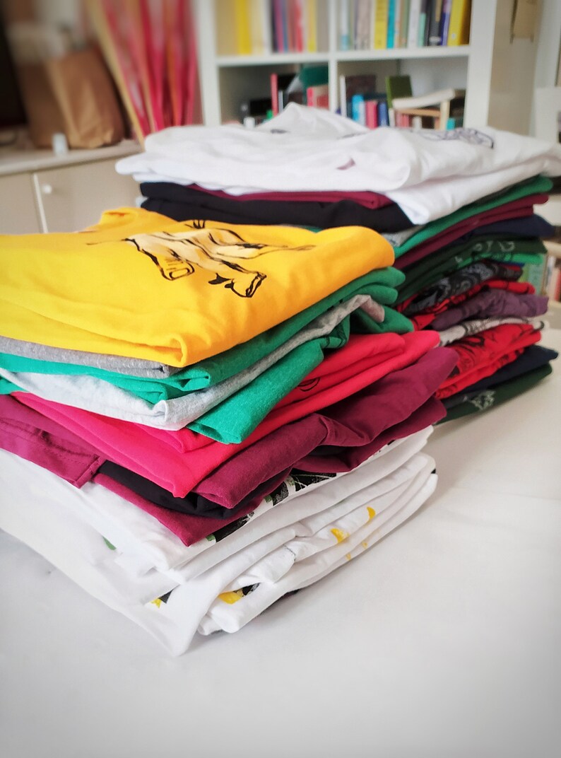 May include: A stack of folded t-shirts in various colors, including white, yellow, green, pink, and burgundy. The shirts are folded neatly and stacked on top of each other.