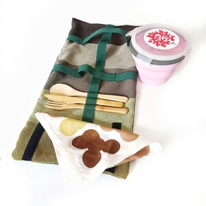 Puede incluir: Un juego de picnic reutilizable con una envoltura de tela a rayas con una correa verde, que contiene cubiertos de bambú. Incluye un recipiente plegable rosa con un diseño de coral y una servilleta estampada.