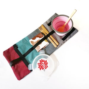 Puede incluir: Un juego de picnic con un cuenco plegable rosa, una cuchara, un tenedor y un cuchillo de madera. El juego incluye una servilleta de tela estampada y un recipiente transparente con un diseño de coral rojo. Los artículos están dispuestos sobre una esterilla rectangular multicolor con correas negras.