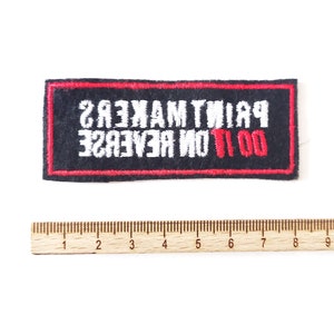 Op de afbeelding: Zwarte geborduurde patch met rode stiksels. De tekst luidt "DO IT ON REVERSE PRINT MAKERS" in wit.