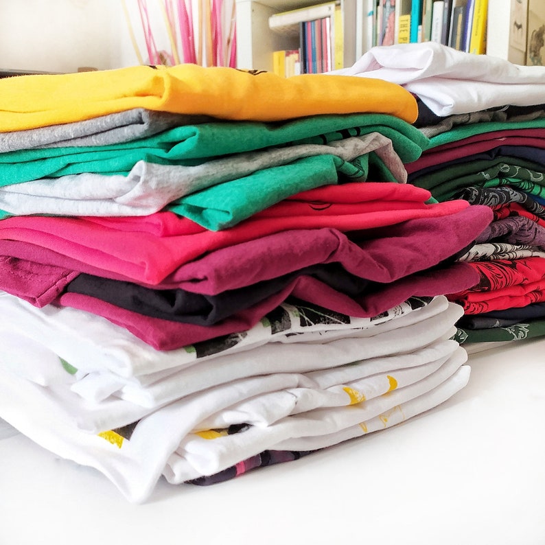 May include: A stack of folded t-shirts in various colors, including yellow, green, red, and white. The shirts are folded neatly and stacked on top of each other.