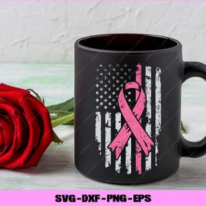 Puede incluir: Taza de caf&eacute; negra con un dise&ntilde;o desgastado de la bandera estadounidense y una cinta rosa, que simboliza la concienciaci&oacute;n sobre el c&aacute;ncer de mama. Una rosa roja descansa junto a la taza. La taza tiene un asa grande.