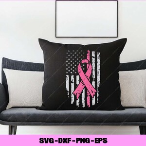 Puede incluir: Coj&iacute;n negro con un dise&ntilde;o de bandera estadounidense desgastada y una cinta rosa. El coj&iacute;n est&aacute; sobre un sof&aacute; gris con dos cojines blancos. La imagen incluye un cuadro con marco negro y una planta.