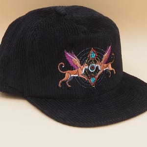 K&ouml;nnte beinhalten: Schwarze Cord-Baseballkappe mit einem gestickten Design. Das Design zeigt zwei gefl&uuml;gelte Kreaturen mit orangefarbenen K&ouml;rpern und lila Fl&uuml;geln, vor einem geometrischen Muster mit t&uuml;rkisfarbenen und orangefarbenen Akzenten.