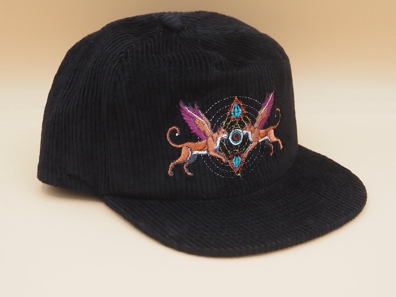 K&ouml;nnte beinhalten: Schwarze Cord-Baseballkappe mit einem gestickten Design. Das Design zeigt zwei gefl&uuml;gelte Kreaturen mit orangefarbenen K&ouml;rpern und lila Fl&uuml;geln, vor einem geometrischen Muster mit t&uuml;rkisfarbenen und orangefarbenen Akzenten.