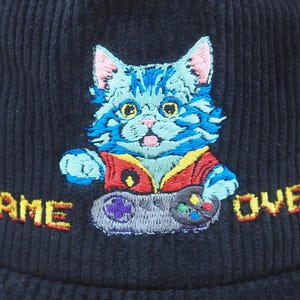May include: Navy blue corduroy hat with an embroidered cartoon cat holding a video game controller. The cat is blue with pink ears and is wearing a red vest. The words "GAME OVER" are embroidered in orange.