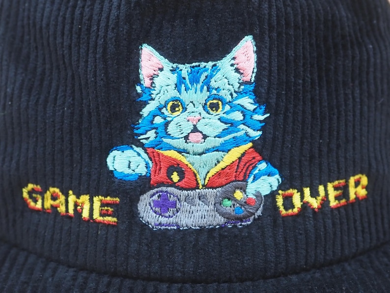May include: Navy blue corduroy hat with an embroidered cartoon cat holding a video game controller. The cat is blue with pink ears and is wearing a red vest. The words "GAME OVER" are embroidered in orange.