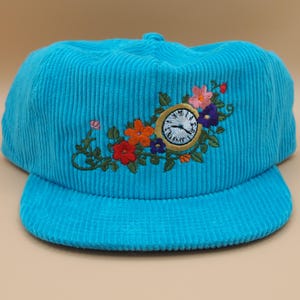 May include: A turquoise corduroy baseball cap with a curved brim. The cap features an embroidered design of a clock surrounded by colorful flowers. The clock face has black hands and a white background.