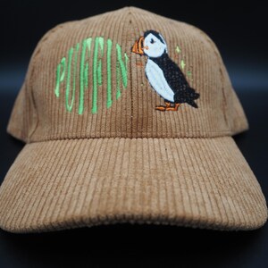K&ouml;nnte beinhalten: Hellbraune Cord-Baseballkappe mit gesticktem Papageientaucher-Design. Das Wort "PUFFIN" ist gr&uuml;n gestickt. Der Papageientaucher ist schwarz, wei&szlig; und orange. Die Kappe hat einen gebogenen Schirm. Ein modisches Accessoire.