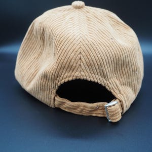 K&ouml;nnte beinhalten: Beige Cord-Baseballkappe mit gebogenem Schirm und verstellbarem Riemen. Die Kappe hat ein sechsteiliges Design mit einem Knopf oben und einem Metallschnallenverschluss. Der Cordstoff hat eine gerippte Textur.