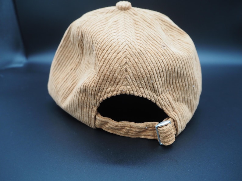 K&ouml;nnte beinhalten: Beige Cord-Baseballkappe mit gebogenem Schirm und verstellbarem Riemen. Die Kappe hat ein sechsteiliges Design mit einem Knopf oben und einem Metallschnallenverschluss. Der Cordstoff hat eine gerippte Textur.
