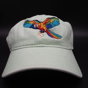 May include: A light green baseball cap with a colorful embroidered parrot design. The parrot has blue, orange, yellow, and red feathers. The cap is made of a soft fabric and has a curved brim.