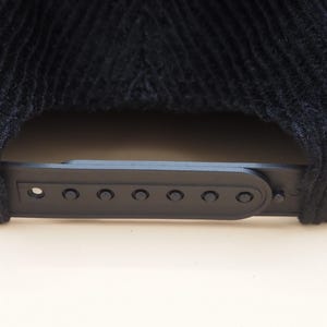 May include: Close-up of a black corduroy baseball cap. The cap features a black plastic adjustable strap with multiple adjustment holes. The corduroy fabric has a ribbed texture.