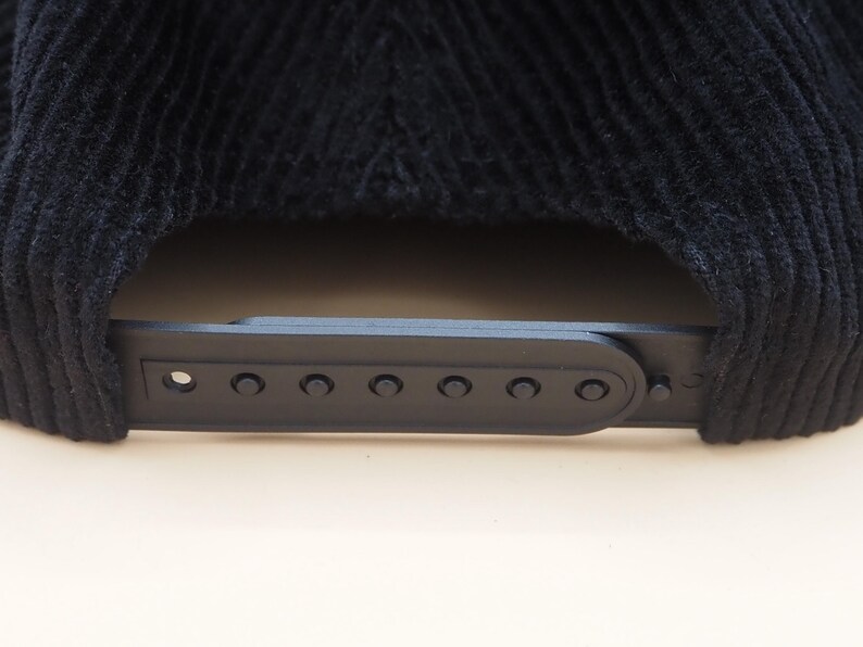 May include: Close-up of a black corduroy baseball cap. The cap features a black plastic adjustable strap with multiple adjustment holes. The corduroy fabric has a ribbed texture.