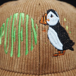 K&ouml;nnte beinhalten: Braune Cord-Baseballkappe mit gr&uuml;n gesticktem Schriftzug &bdquo;PUFFIN&ldquo; und einem Papageientaucher-Design. Der Papageientaucher hat eine wei&szlig;e Brust, einen schwarzen R&uuml;cken, einen orangefarbenen Schnabel und F&uuml;&szlig;e. Ein modisches Accessoire.