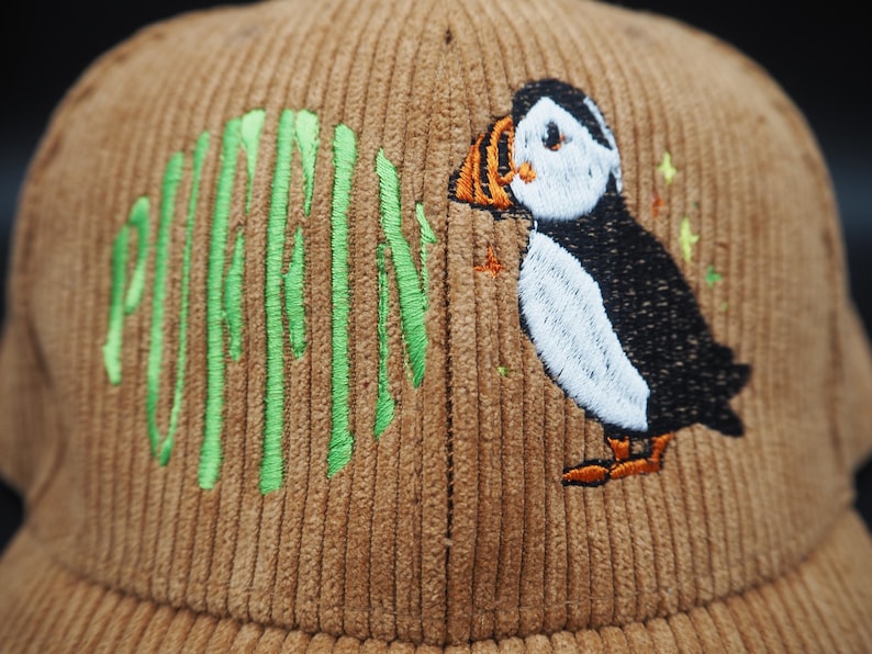 K&ouml;nnte beinhalten: Braune Cord-Baseballkappe mit gr&uuml;n gesticktem Schriftzug &bdquo;PUFFIN&ldquo; und einem Papageientaucher-Design. Der Papageientaucher hat eine wei&szlig;e Brust, einen schwarzen R&uuml;cken, einen orangefarbenen Schnabel und F&uuml;&szlig;e. Ein modisches Accessoire.