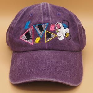 May include: Purple baseball cap with embroidered design. The design includes colorful triangles with birds, a skull, and geometric shapes. The cap is made of a soft, textured fabric.