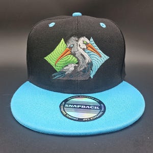 May include: Black snapback cap with a light blue brim and top button. The cap features an embroidered design of a gray bird with orange beak and green eyes, set within a diamond-shaped pattern. The text "SNAPBACK" is visible on the brim.