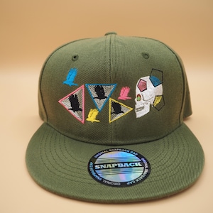 May include: Olive green snapback cap with embroidered designs. Features a skull and geometric shapes, along with triangles with bird silhouettes. The brim has a holographic sticker that says "SNAPBACK".