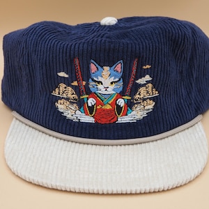 May include: Navy blue corduroy cap with a white brim. The cap features an embroidered design of a cat in a samurai outfit, holding two swords. The cat has blue and white fur and is wearing a red and green outfit.