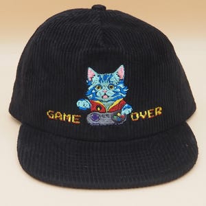 May include: Black corduroy baseball cap with an embroidered design of a blue cat holding a video game controller. The text "GAME OVER" is embroidered in yellow. The cap has a curved brim and a button on top.