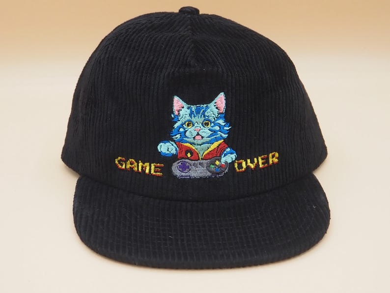 May include: Black corduroy baseball cap with an embroidered design of a blue cat holding a video game controller. The text "GAME OVER" is embroidered in yellow. The cap has a curved brim and a button on top.