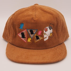 May include: A brown corduroy baseball cap with a curved brim. The cap features embroidered designs including a skull, triangles with birds, and colorful accents. The cap is a stylish accessory.