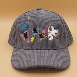 May include: Gray corduroy baseball cap with embroidered designs. Features a skull, birds, and geometric shapes in various colors. The cap has a curved brim and a button on top.